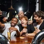 happy multicultural men taking selfie on smartphone while drinking beer in bar, bachelor party