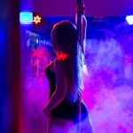 Pole dance. Slender sexy woman dancing in the interior of a nightclub with a colored background and smoke.
