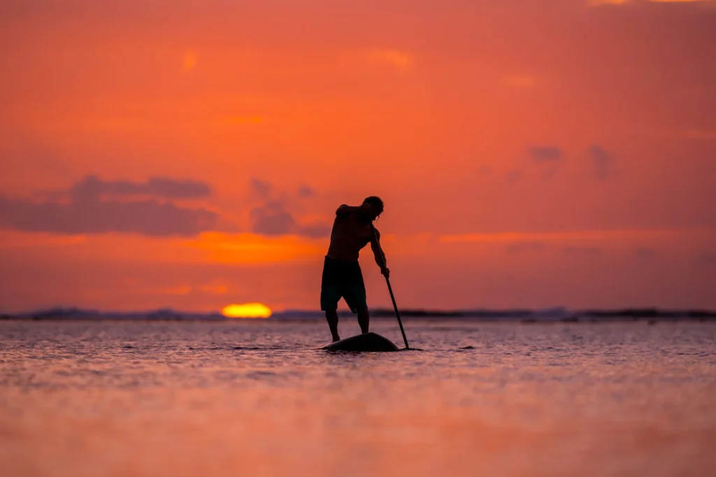 Surfeur en paddle board (S.U.P.) dans l'océan sur fond d'un grand disque de soleil couchant.