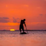 surfer rides by paddle board (S.U.P.) in the ocean against the background of a large disk of the setting sun
